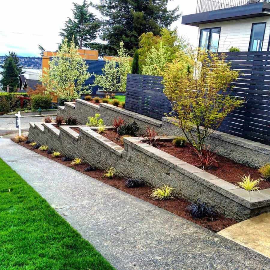 Stepped concrete retaining walls with modern landscaping, featuring trees, shrubs, and mulch, bordered by a clean walkway and wooden privacy fencing