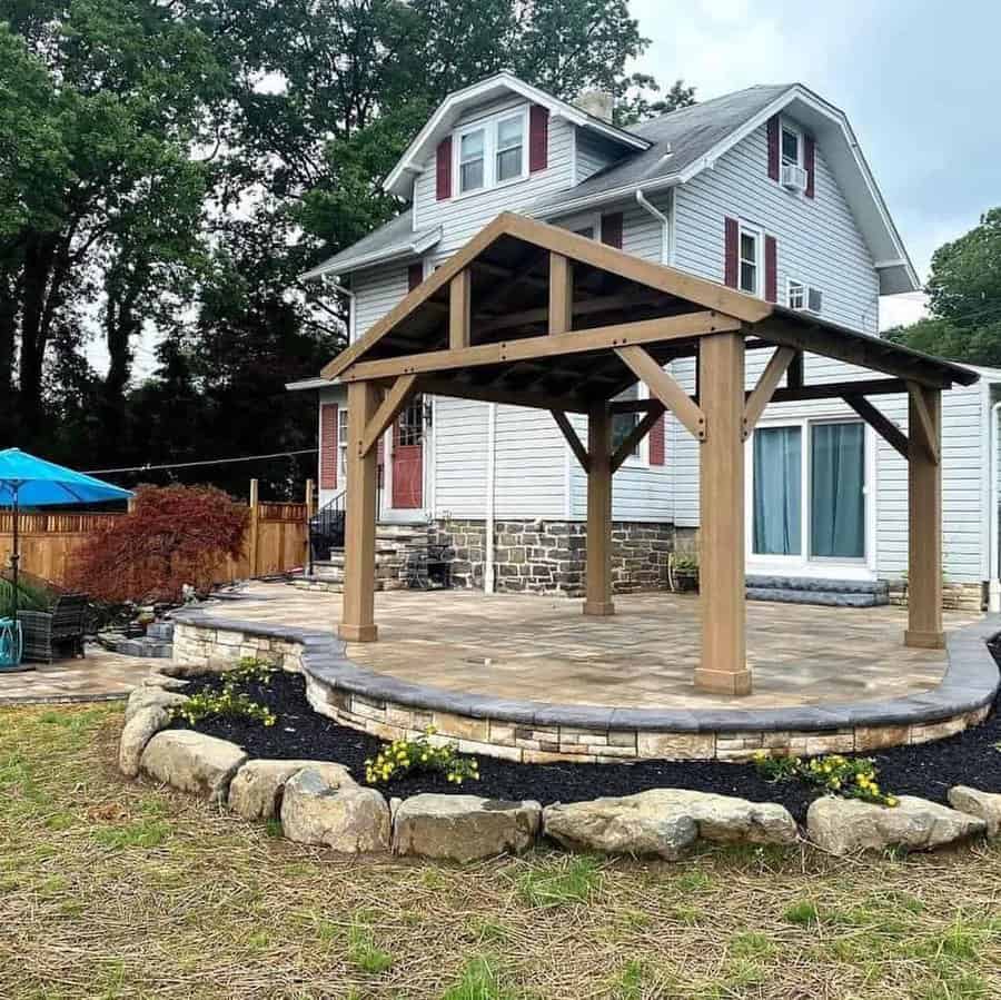 Backyard patio with a wooden pergola, raised stone retaining wall, and neatly landscaped garden featuring mulch and flowers, set against a traditional house