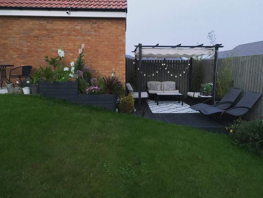 Cozy sloped backyard with a lush lawn, raised planters, a pergola-covered seating area adorned with string lights, and stylish lounge chairs