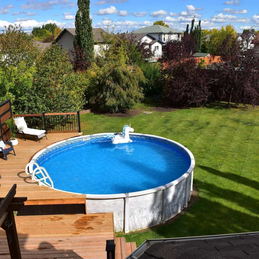 Above-ground pool with blue water, a unicorn float, and wooden deck, surrounded by lush green grass and trees