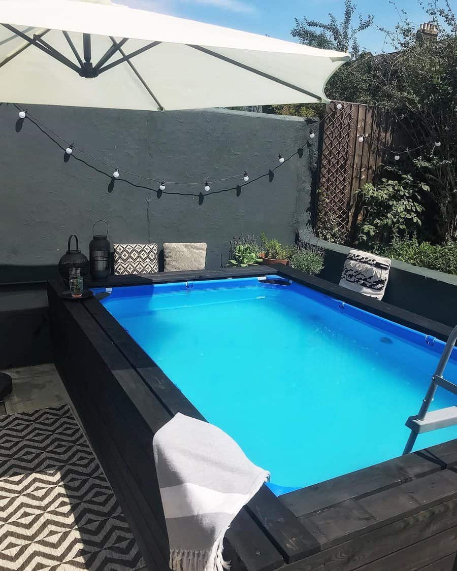 Cozy above-ground pool with a black wooden frame, soft seating, and string lights, creating a stylish and intimate backyard retreat