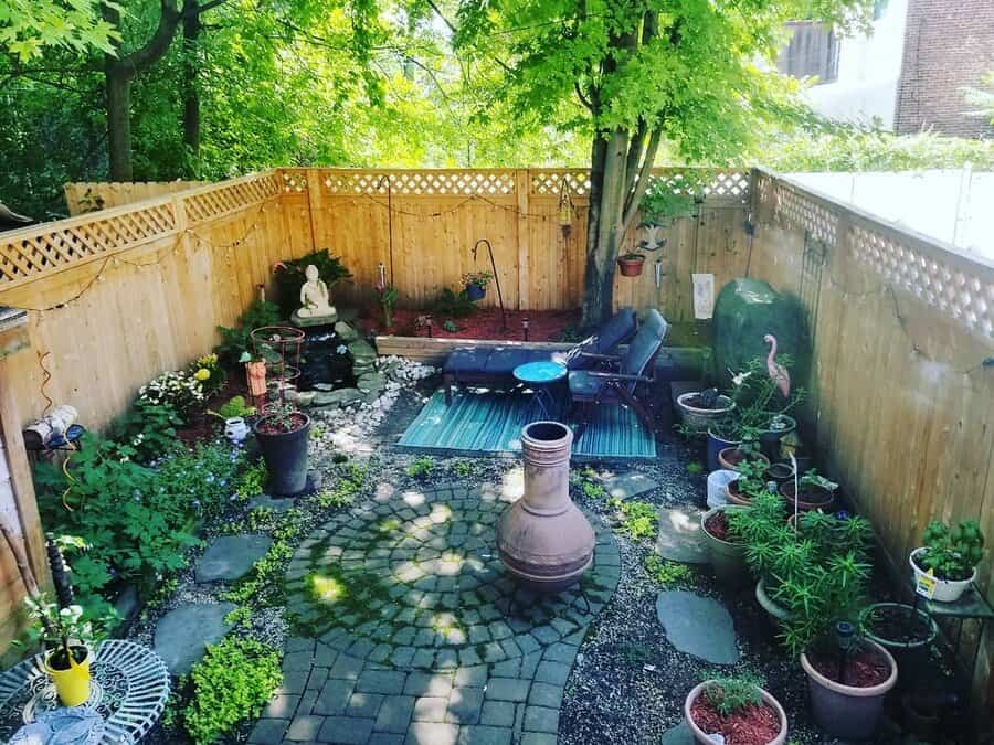 A cozy backyard garden with a seating area, potted plants, a small fountain, a statue, and a chiminea surrounded by a wooden fence