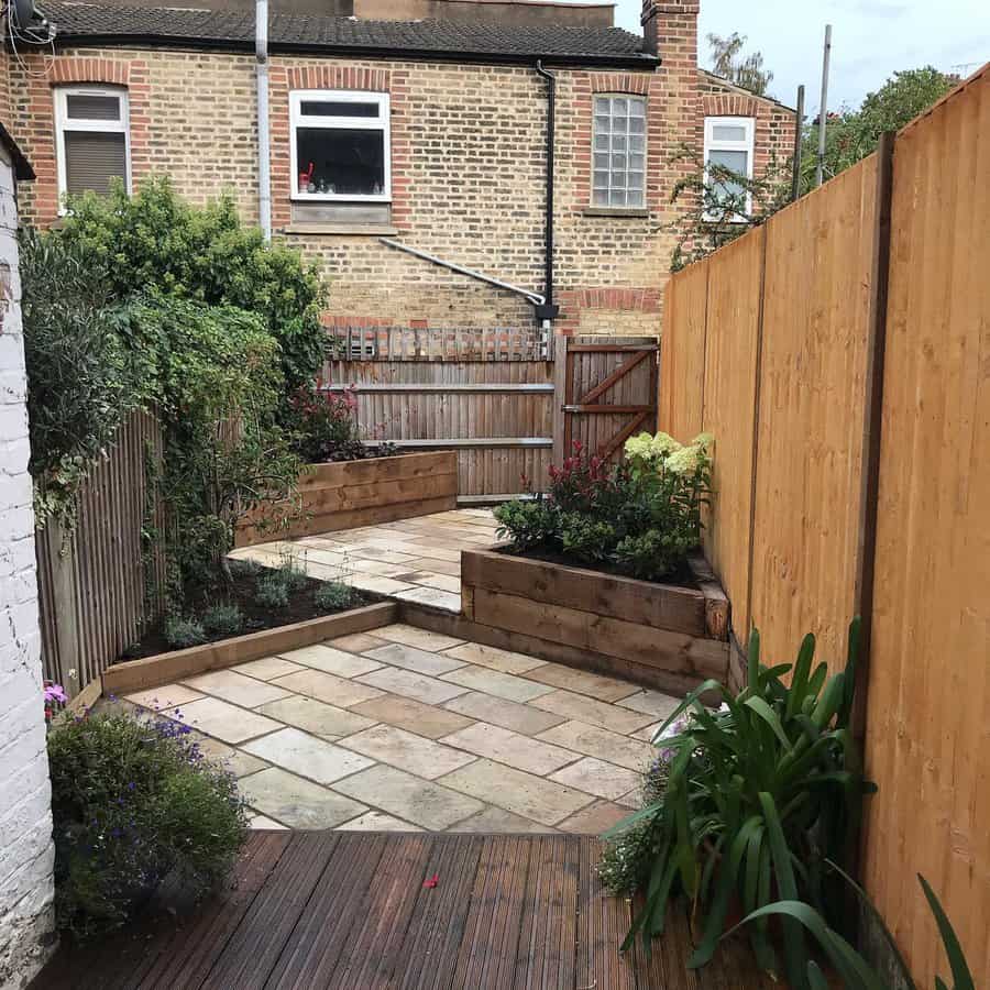 A narrow backyard with a paved path, wooden planters, and a tall fence; brick building is visible in the background