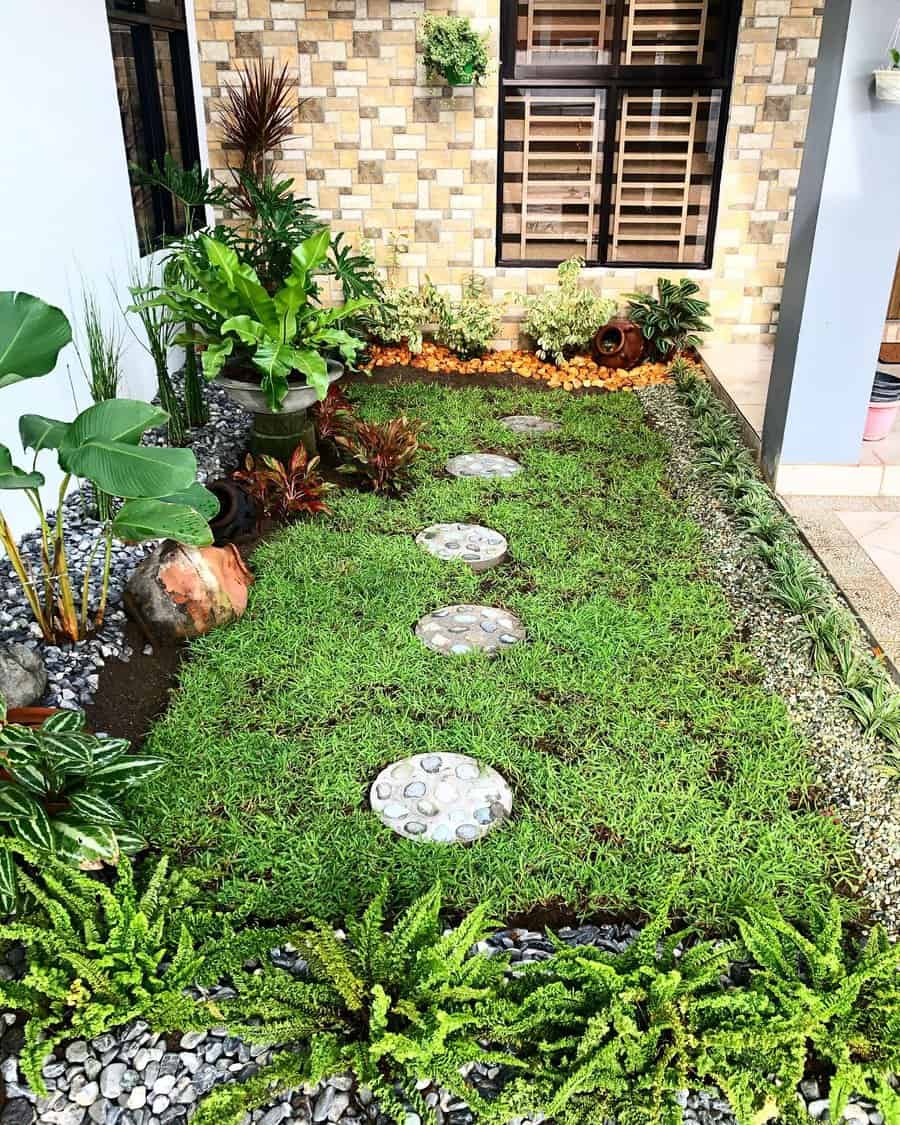 Small garden with green grass, stepping stones, various plants, and a stone border, adjacent to a brick wall and window