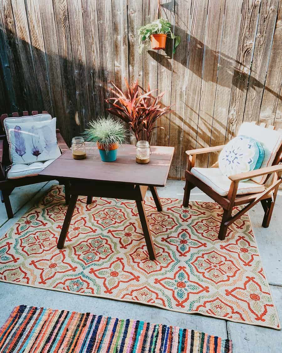 Cozy outdoor patio with two chairs, a wooden table, patterned rugs, potted plants, and a wooden fence background