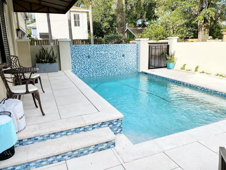 Outdoor pool with blue mosaic tiles, surrounded by a patio with chairs and plants, adjacent to a building