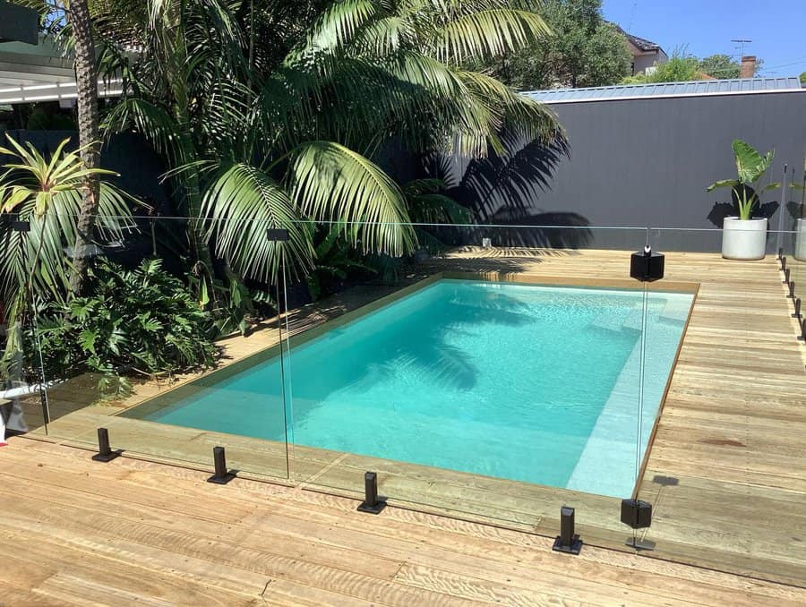 A small rectangular pool with clear glass fencing, surrounded by wooden decking and tropical plants