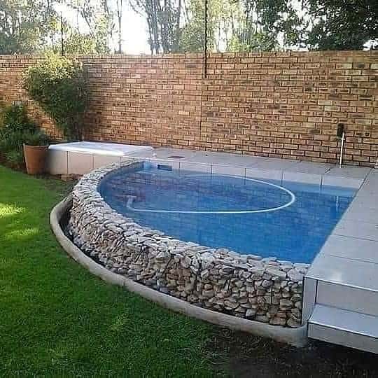 A round elevated swimming pool with a stone base, surrounded by green grass and a brick wall in the background