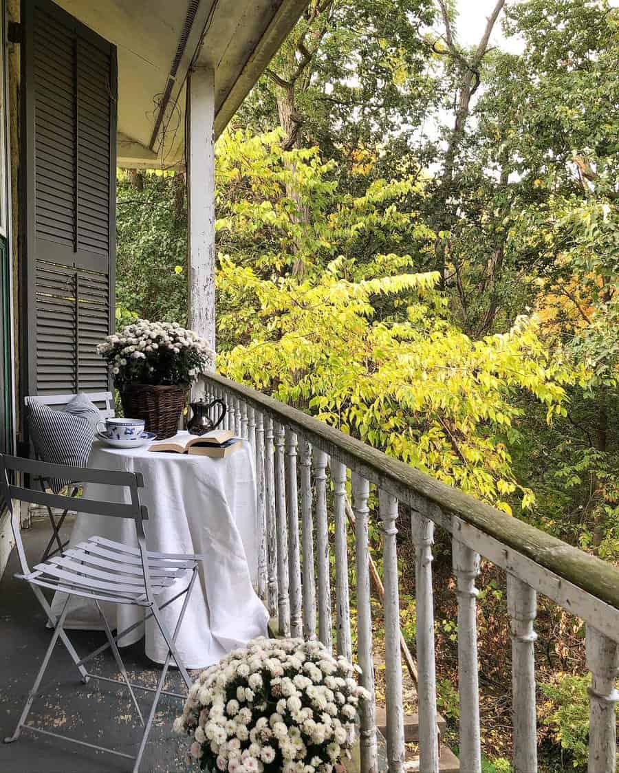 A cozy balcony with a table, open book, teacup, and flowers, surrounded by lush green and yellow trees with a folding chair nearby