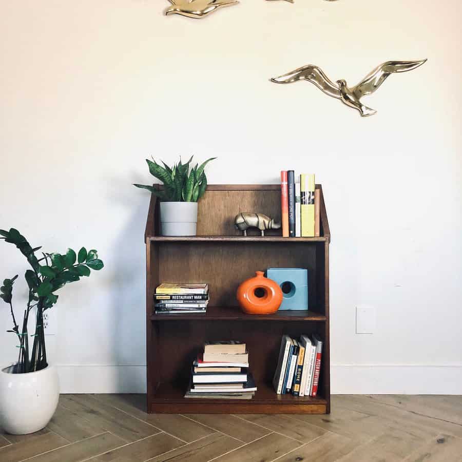 Stylish wooden bookshelf with books, plants, and modern decor, set against a white wall with gold bird sculptures and a light wood floor.