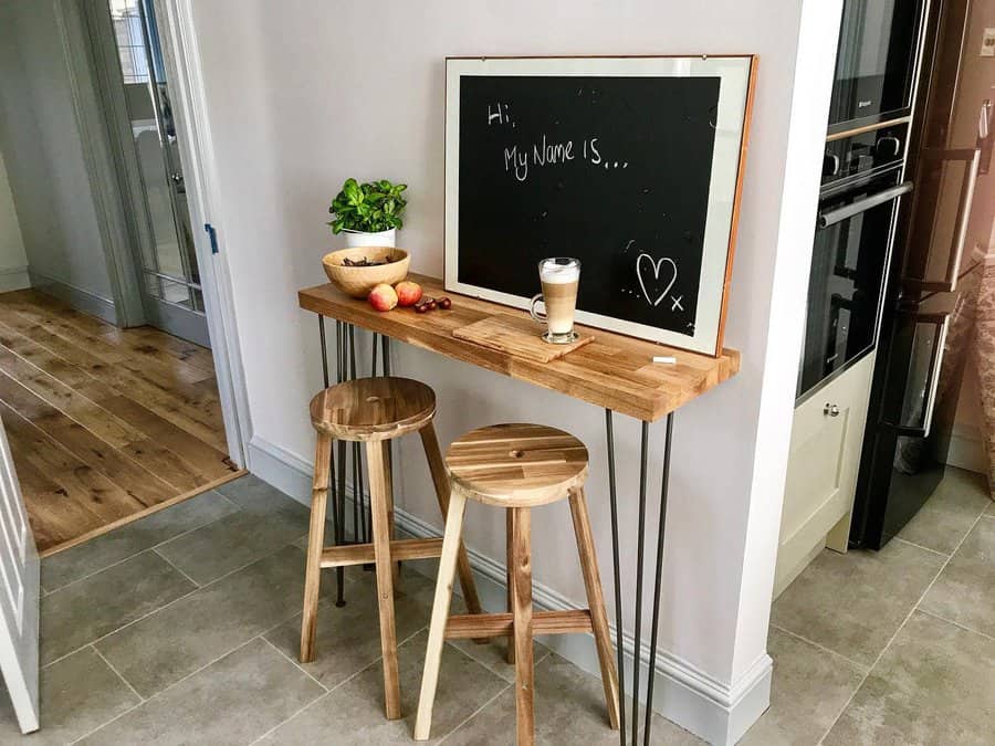 Wooden bar table with two stools, chalkboard, plant, fruit bowl, and a coffee cup on top; light-filled room with tiled floor