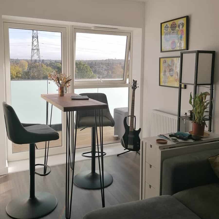 Compact breakfast bar setup in a modern apartment, featuring a slim wooden table with metal legs, sleek black barstools, and a scenic window view