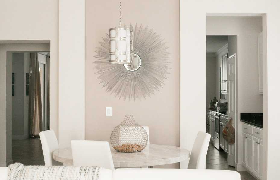 Modern dining area with a round table, white chairs, decorative vase, sunburst wall art, and a hanging light fixture; kitchen visible