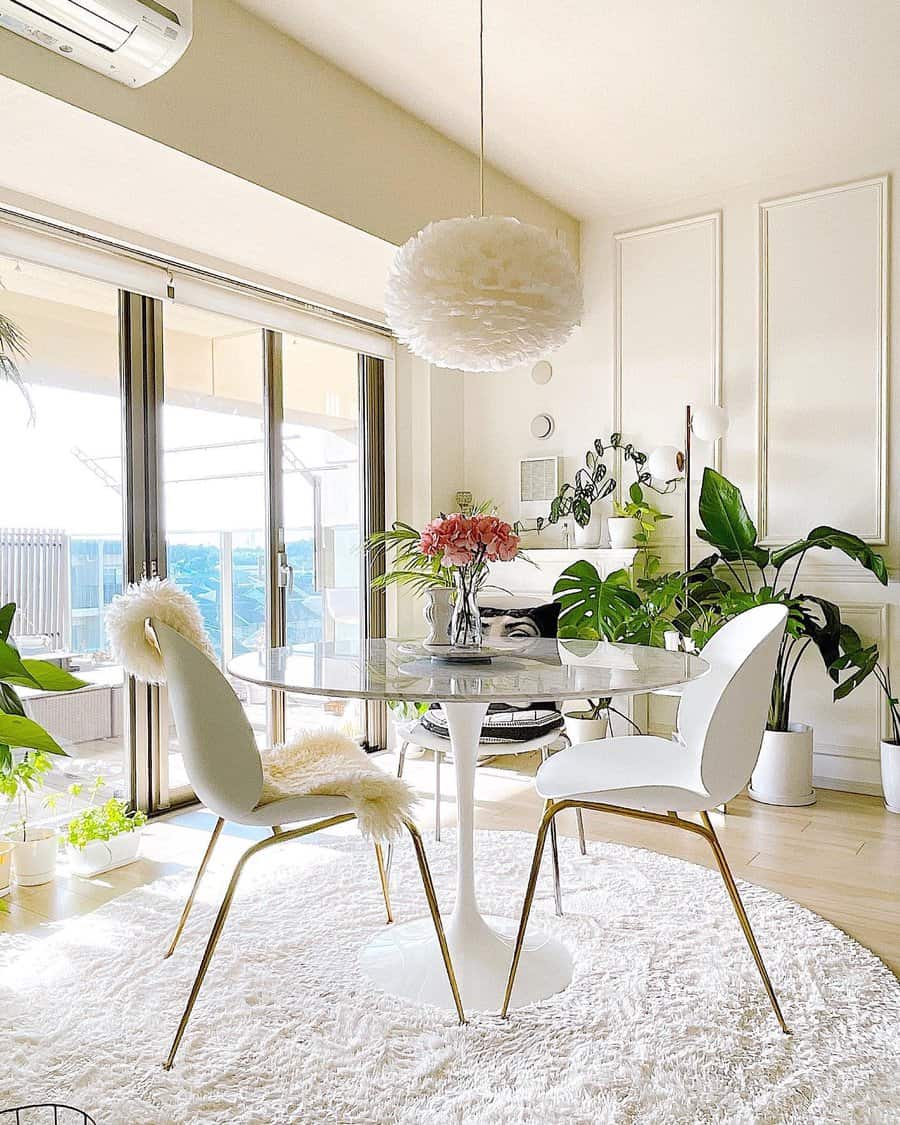 Bright dining room with a round table, two chairs, a fluffy pendant light, and plants; floor-to-ceiling windows overlook a balcony