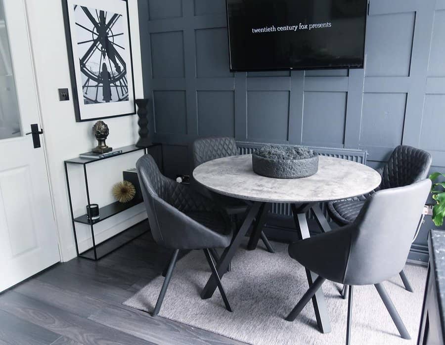 Modern dining area with a round marble table, four quilted black chairs, a wall-mounted TV, and minimalist decor in a gray-toned room