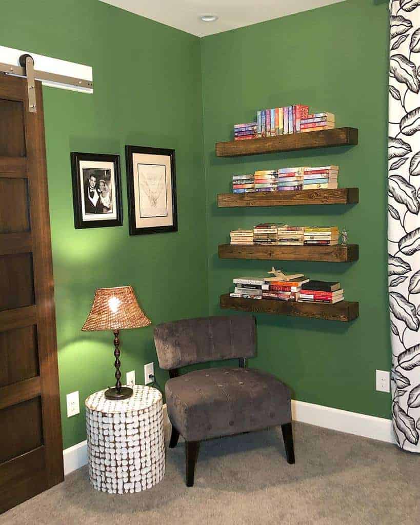 Cozy reading nook with a gray chair, round side table, and four shelves of books on a green wall, next to framed pictures