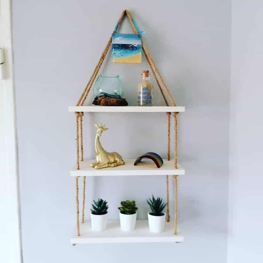 Triangle rope shelf with beach photo, glass bottle, gold giraffe, rainbow ornament, and three small potted plants on a gray wall