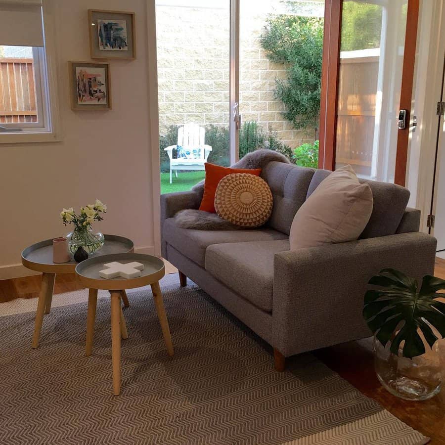 Cozy small living room with a gray loveseat, textured pillows, round nesting tables, and sliding glass doors opening to a lush outdoor space