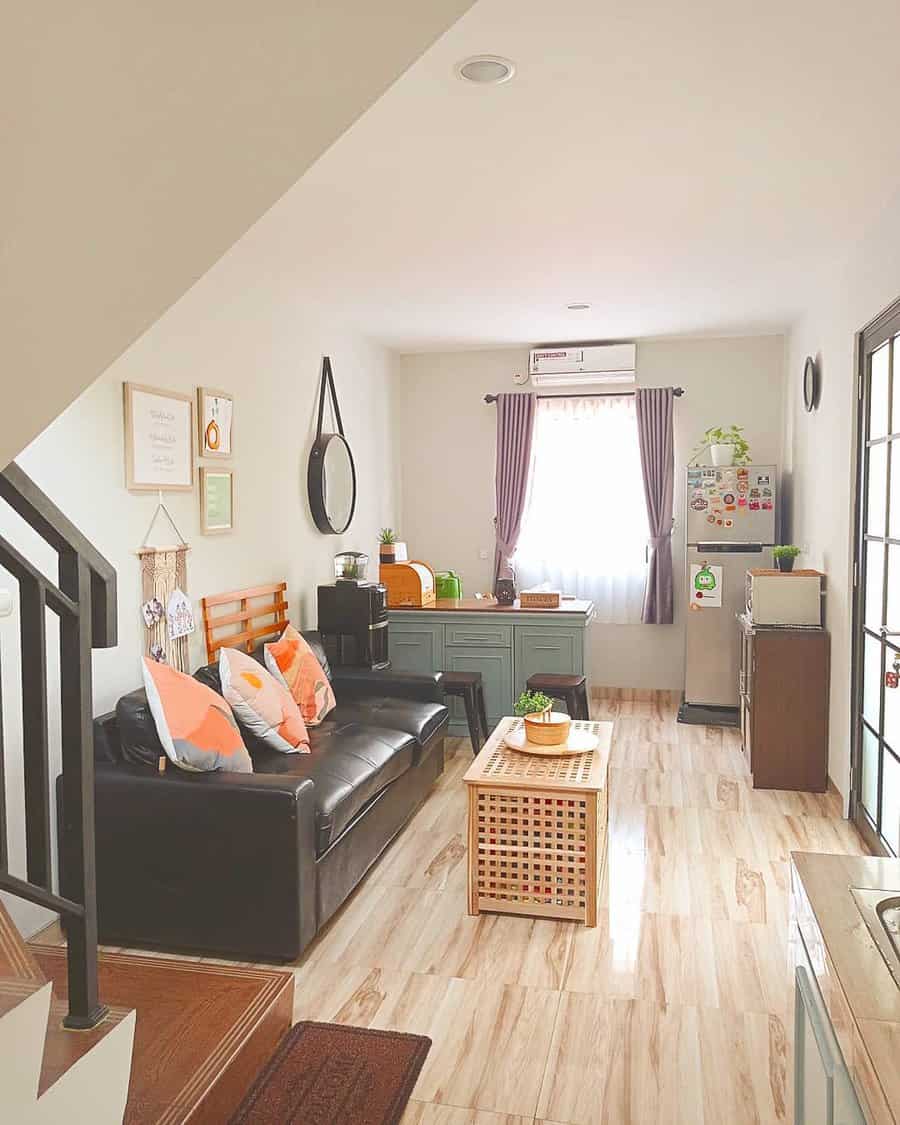 Compact and cozy living room with a black leather sofa, pastel accent pillows, wooden furniture, and soft natural light for a warm and inviting space