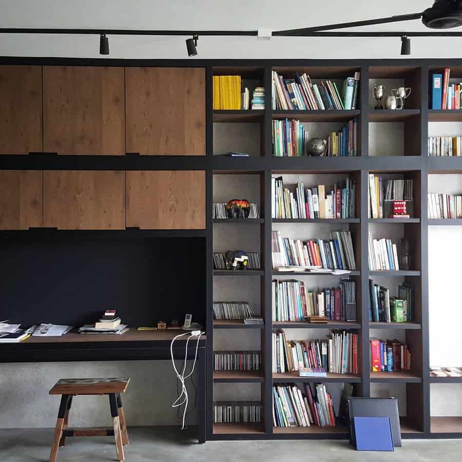 A modern home office with a wooden desk, shelves filled with books, closed cabinets above, and a wooden stool in front