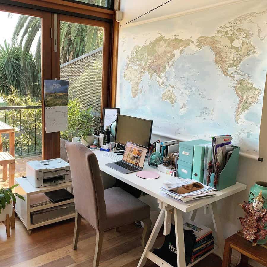 Cozy home office with a world map, desk, computer, and office supplies, large window view of greenery and a balcony with a table