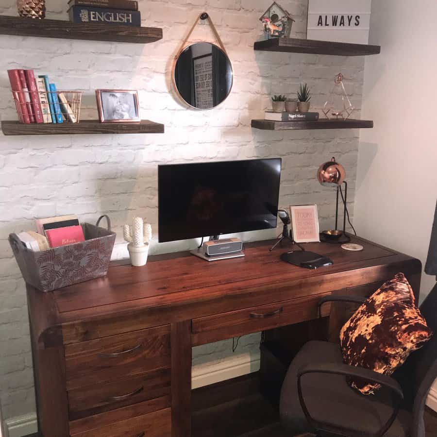 Cozy home office with a wooden desk, computer, and decor on shelves; a small mirror and plants add charm to the brick wall backdrop