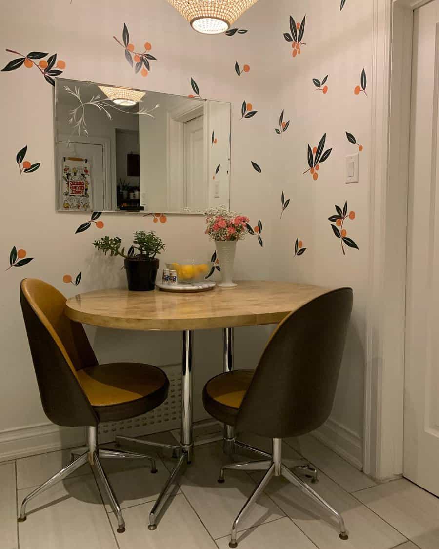 Small dining nook with a round wooden table, two brown chairs, floral wallpaper, a mirror, and small decorative plants