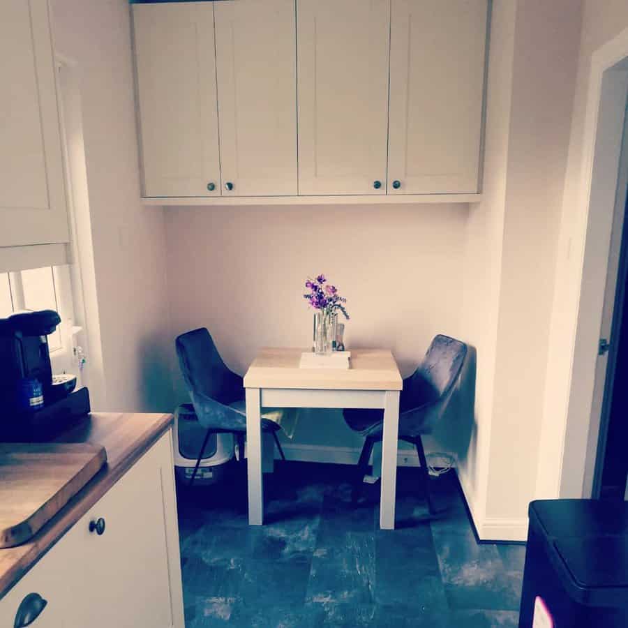 Cozy corner kitchen nook with a small table, two chairs, a vase of flowers, and white cabinets above