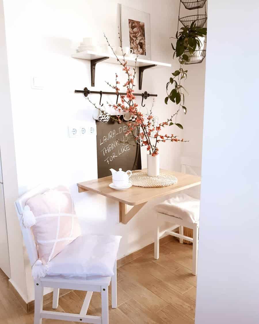 Cozy corner with a small wooden table, white chairs, pink cushions, cherry blossom branches in a vase, and wall shelves with decor