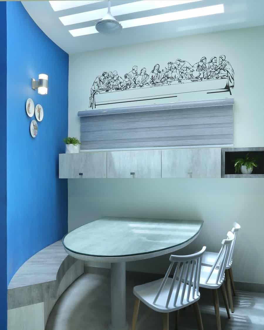 Modern dining area with a blue accent wall, circular table, three chairs, and a wall sketch of "The Last Supper" above cabinets