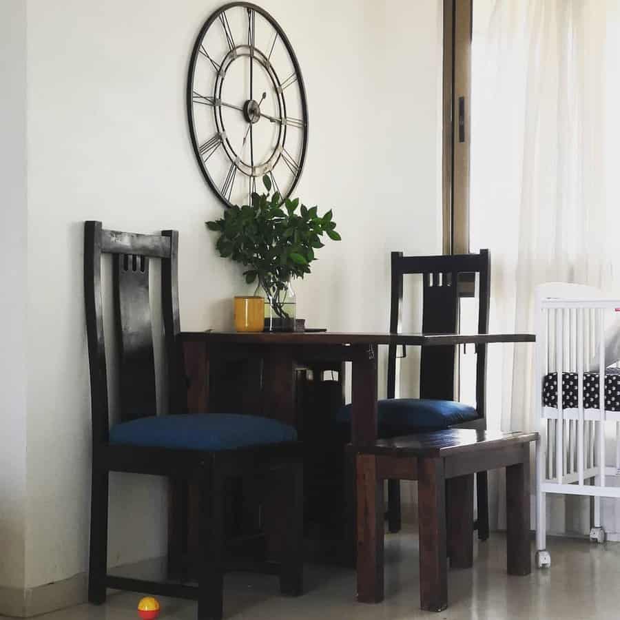 Wooden table set with two chairs under a large wall clock, with a small plant and mug on the table, a crib, and curtain nearby