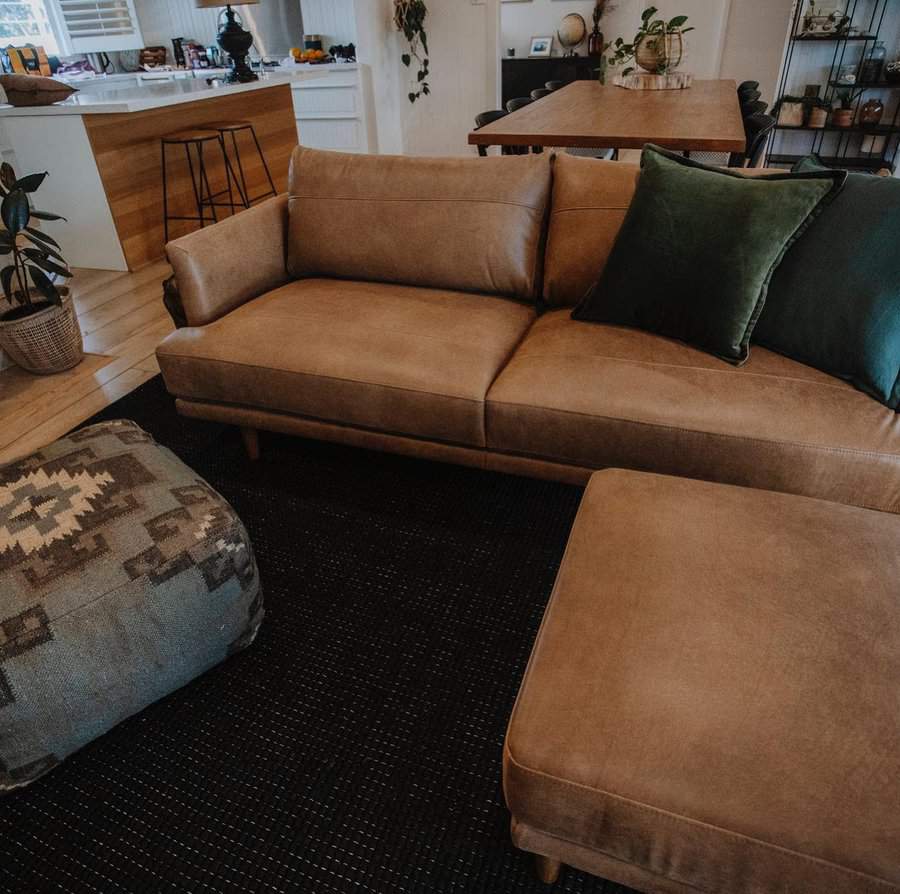 Living room with a tan leather couch, green pillows, ottoman, and a dining table