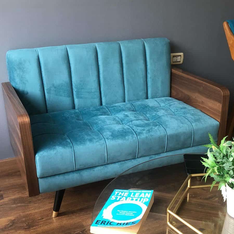 Stylish tiny living room with a retro-inspired teal velvet sofa, dark wooden armrests, a glass coffee table, and a modern accent book for a chic touch