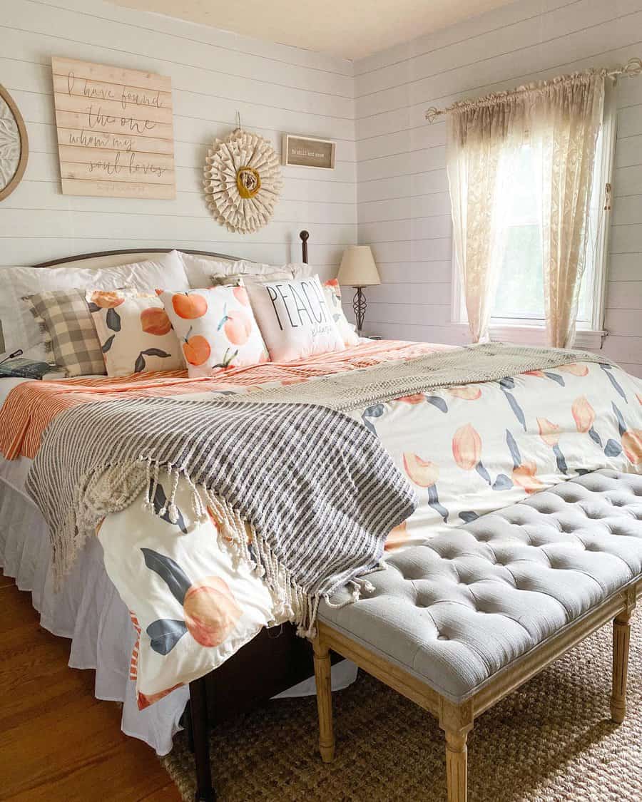 Bedroom with a floral bedspread, decorative pillows, and a tufted bench; beige curtains allow light in, with wall art above the bed