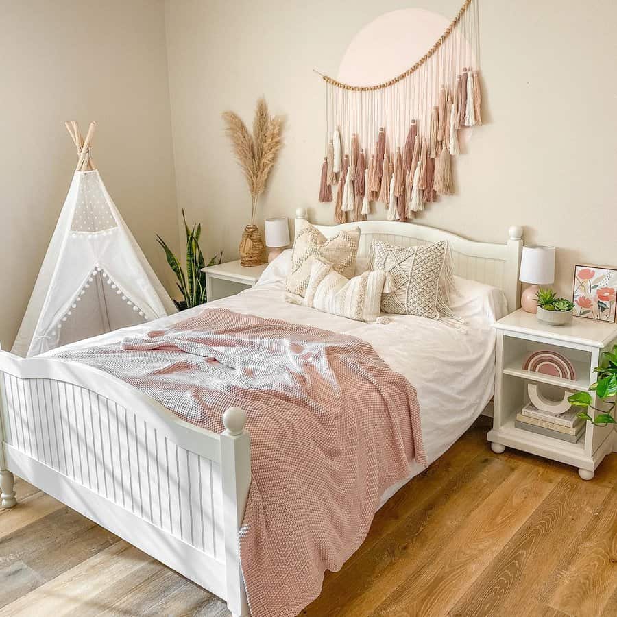 Cozy bedroom with a white bed, pink and cream bedding, tassel wall decor, teepee, and plants on a wooden floor