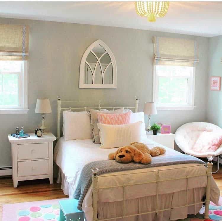 Bright bedroom with a white bed, plush dog toy, nightstand, and arched mirror. Soft pastel accents and sunlight fill the space