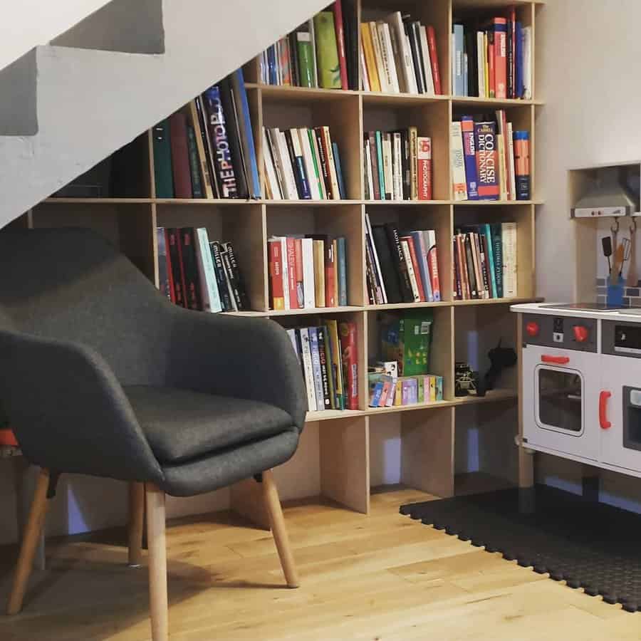 Space-saving under-stair bookshelf filled with books, a cozy reading chair, and a children's play kitchen set on a wooden floor.