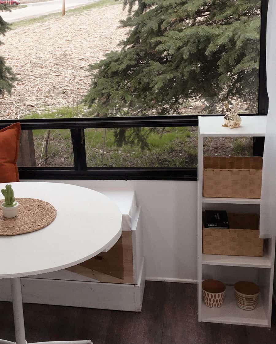 Cozy RV interior with a round table, cactus plant, orange pillow, window view of trees, and shelving unit with baskets