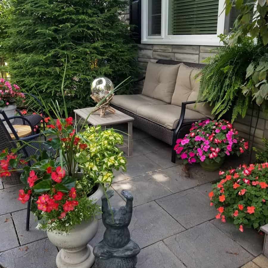 Cozy backyard patio with square stone pavers, a cushioned wicker sofa, lush greenery, and vibrant flowers for a charming outdoor retreat