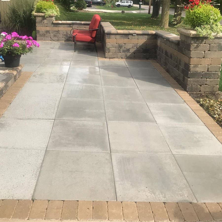 Patio with large gray tiles, a brick border, a single red cushioned chair, and potted flowers in a peaceful garden setting