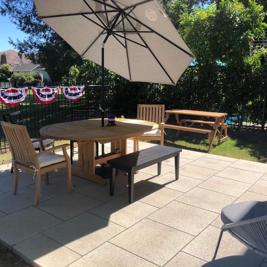 Sunny backyard patio with large square pavers, a wooden dining set, a shaded umbrella, and a picnic table for a relaxed outdoor gathering space