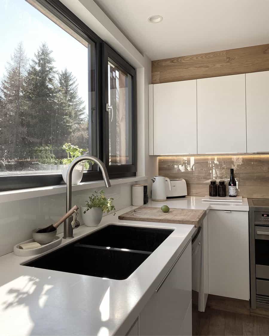 Modern kitchen with white cabinets, black sink, and large window where sunlight illuminates plants, a cutting board, and kitchen items