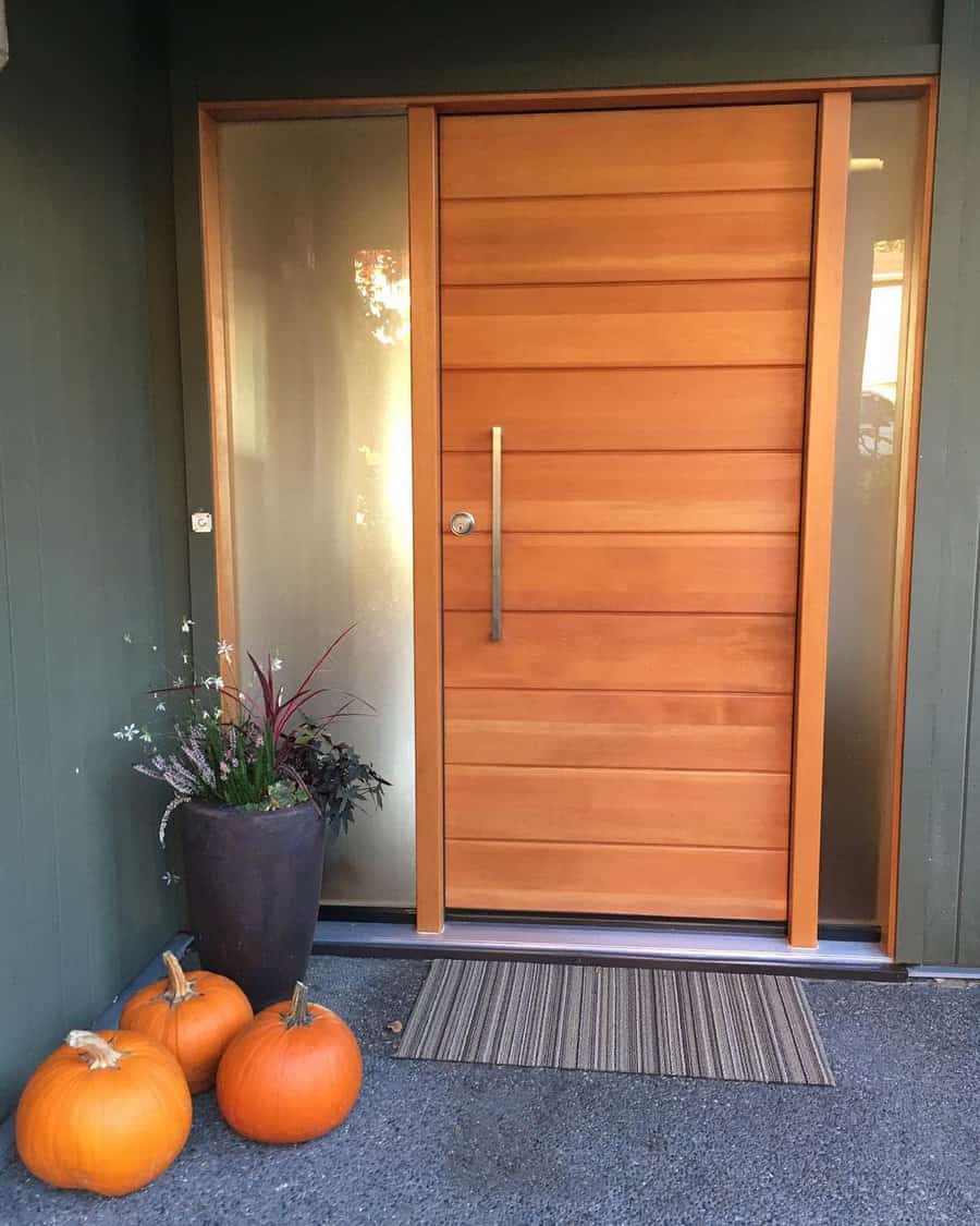 A modern wooden front door with a vertical handle, flanked by frosted glass panels; pumpkins and potted plants decorate the entrance