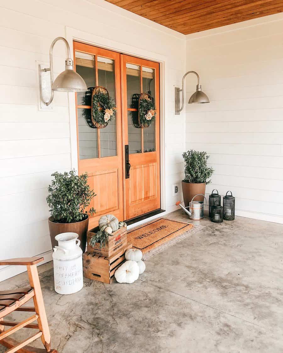 Front porch with wooden doors, fall wreaths, potted plants, lanterns, pumpkins, and a welcome mat; rustic decor and white siding