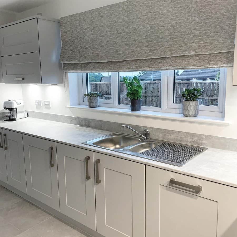 Modern kitchen with light grey cabinets, a double stainless steel sink, potted plants on the windowsill, and a large window with a grey blind