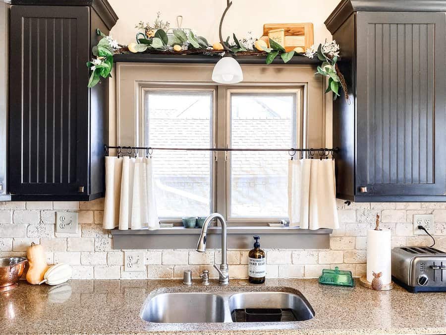 Rustic kitchen with a stainless steel double sink, high-arch faucet, dark wood cabinets, stone backsplash, and farmhouse-style window decor