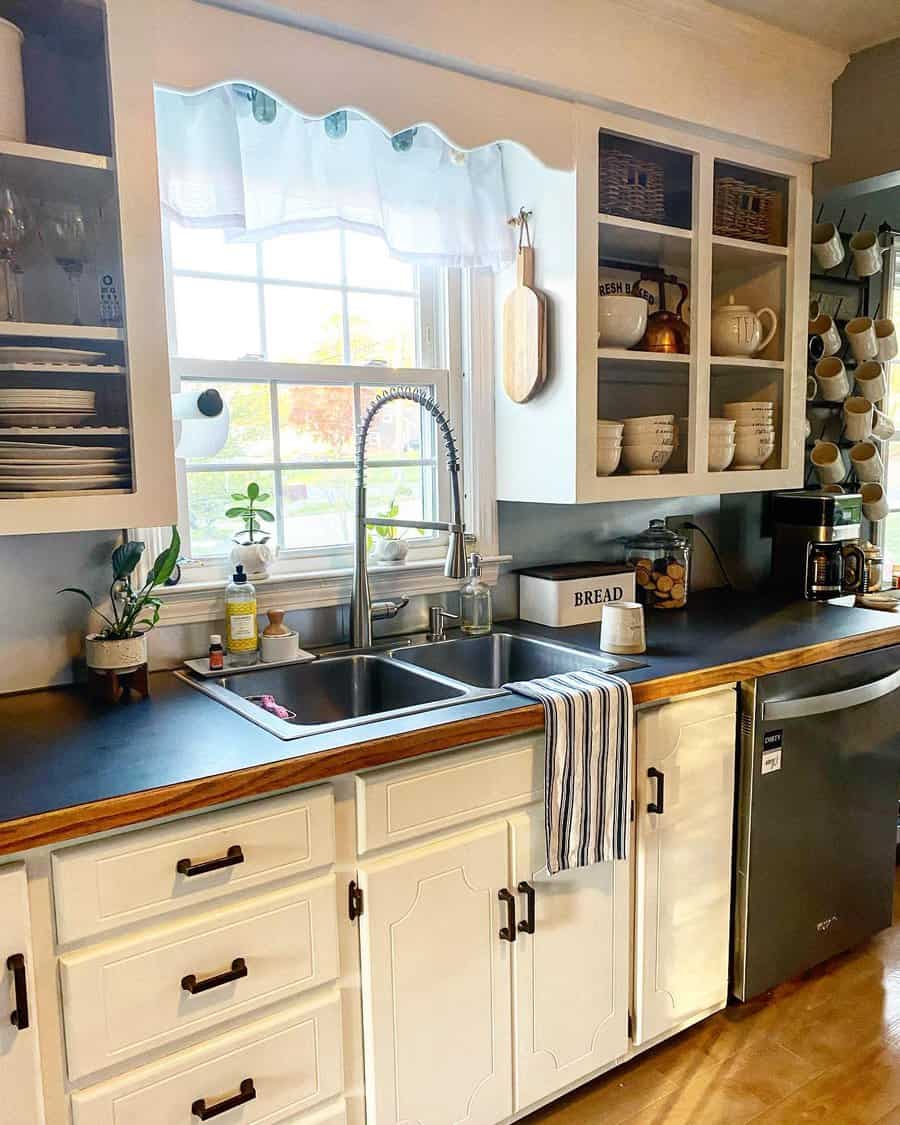 Cozy farmhouse kitchen with a stainless steel double sink, high-arch spring faucet, black countertops, open shelving, and charming window decor