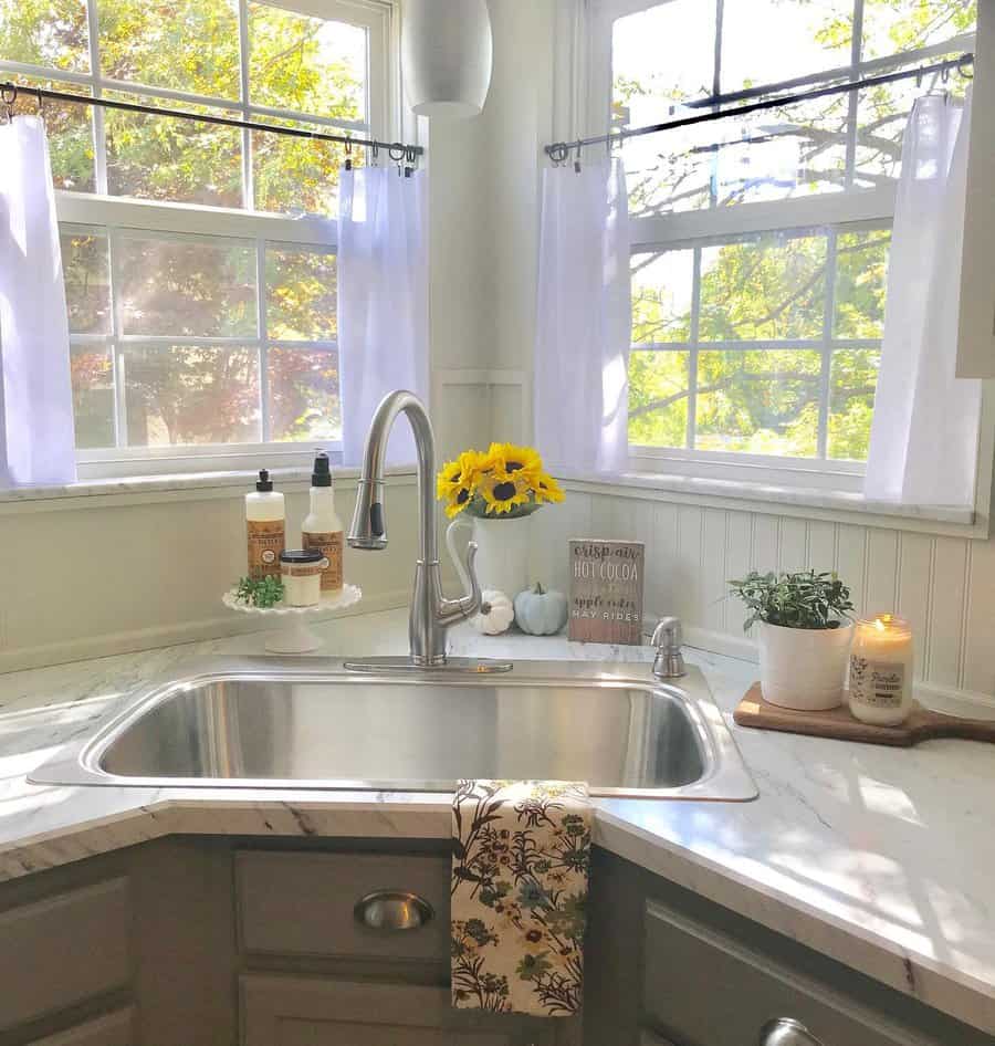 Bright farmhouse kitchen with a corner stainless steel sink, marble countertops, cozy window curtains, and fresh sunflowers adding a cheerful touch