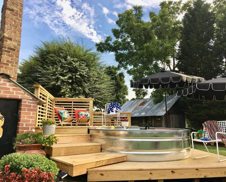 Rustic stock tank pool with a wooden deck, colorful seating, and chic black umbrellas, creating a cozy and stylish outdoor retreat