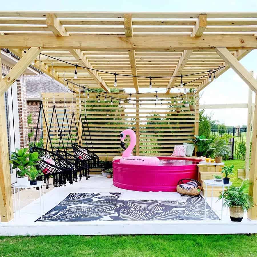 Vibrant pink stock tank pool under a wooden pergola with boho hanging chairs, lush greenery, and string lights for a fun backyard oasis